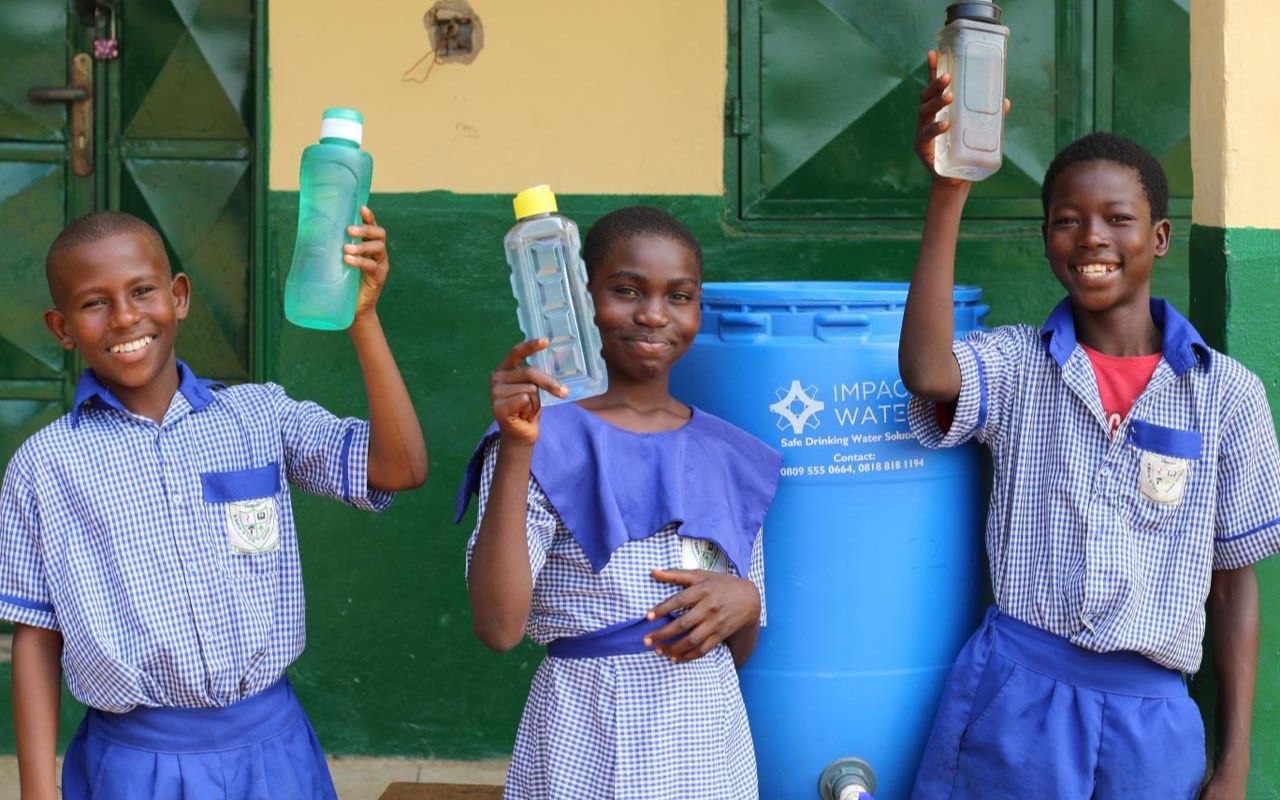 Schulkinder mit Wasserflaschen in der Hand vor einem Wassertank in dem Wasser aufbereitet wird